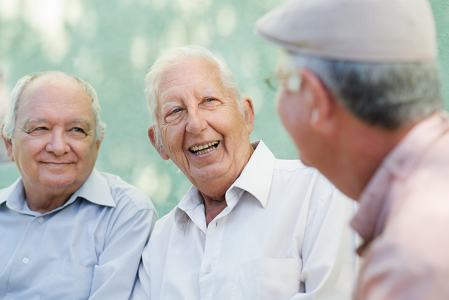 Active retirement group of three old male friends talking and laughing on bench in public park